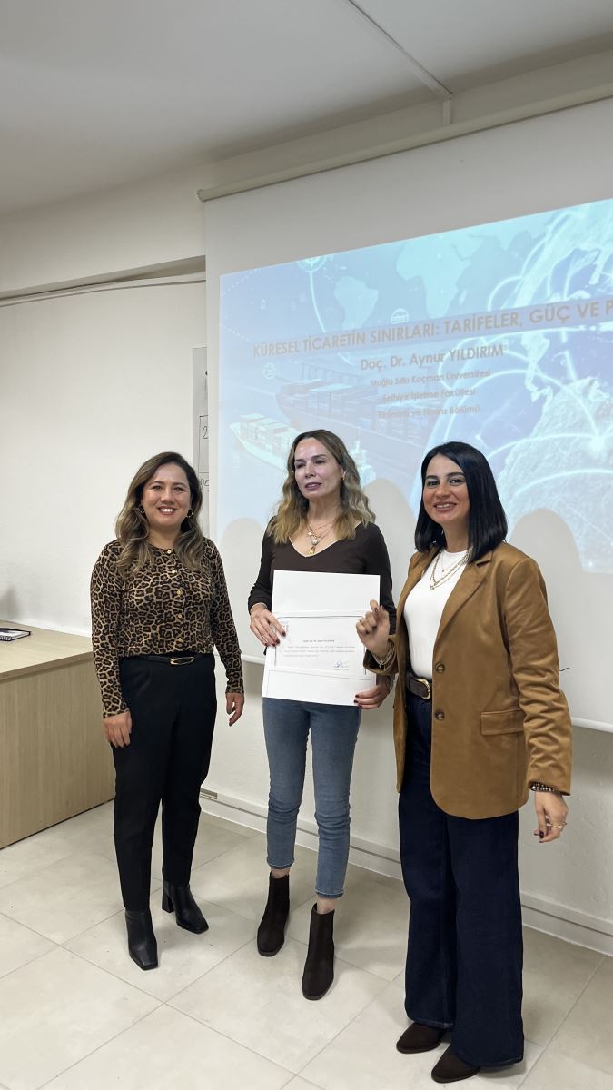 Üç kadın, sınıf ortamında bir projeksiyon perdesinin önünde yan yana duruyor. Ortadaki kadın elinde bir sertifika tutuyor. Kadınların hepsi gülümser durumda. Arka planda projeksiyon perdesine yansıtılmış bir sunum bulunuyor. Sunumun arka planı dünya haritası, bir yük gemisi ve dijital çizgilerden oluşuyor. Sunum başlığında “Küresel Ticaretin Sınırları: Tarifeler, Güç ve Politika” yazısı yer alıyor. Oda sınıf düzeninde, beyaz duvarlı ve sade bir ortam.